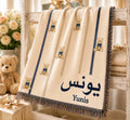 Personalized baby blanket with a teddy bear, navy ribbon, and custom Arabic name, draped over a plush teddy bear.