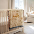 Classic Framed Arabic Calligraphy blanket draped over a white crib, with a plush teddy bear and a brightly lit nursery background.