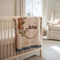Custom baby blanket showcasing a botanical teddy bear and Arabic name, draped over a white crib beside a window.