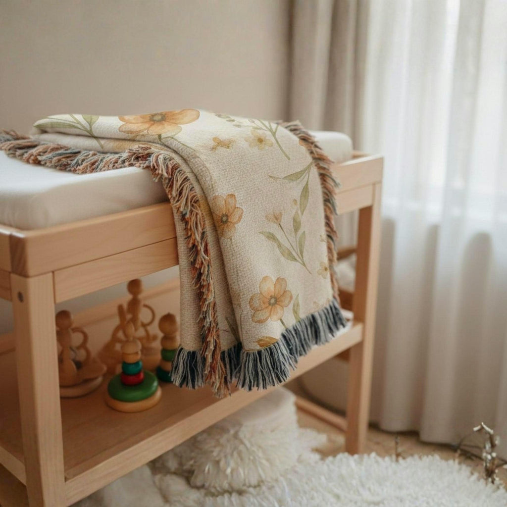 Framed floral pattern baby blanket draped across a padded bench, next to a wooden toy on the floor in a softly lit room, highlighting its tasseled fringe and layered appearance.
