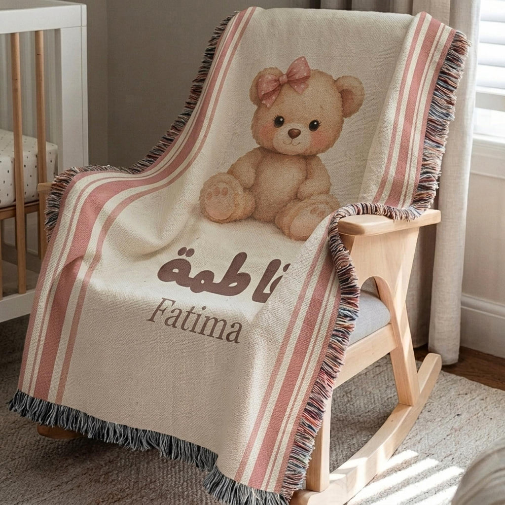 Vintage bear patterned baby blanket draped over a wooden rocking chair, highlighting the blush pink stripes, teddy bear illustration, and fringed edges.
